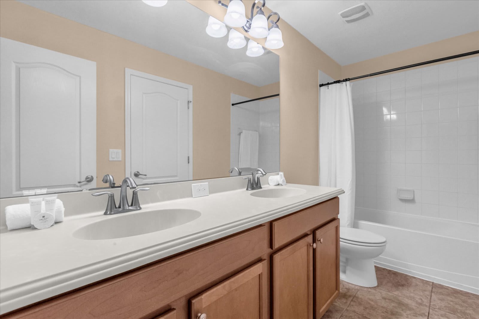 Bathroom with a light wood double vanity, two white sinks, a large mirror with decorative lighting, and a shower-tub combo with white subway tiles and a white shower curtain.