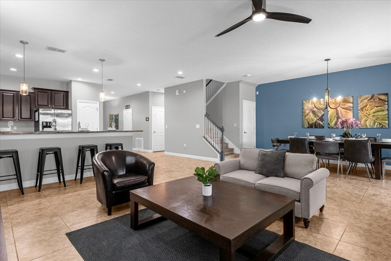 Spacious open-concept living area featuring a light gray sofa, a dark wood coffee table, and a brown leather armchair on a dark rug. The room connects to a kitchen bar, dining area with a blue accent wall, and a staircase.