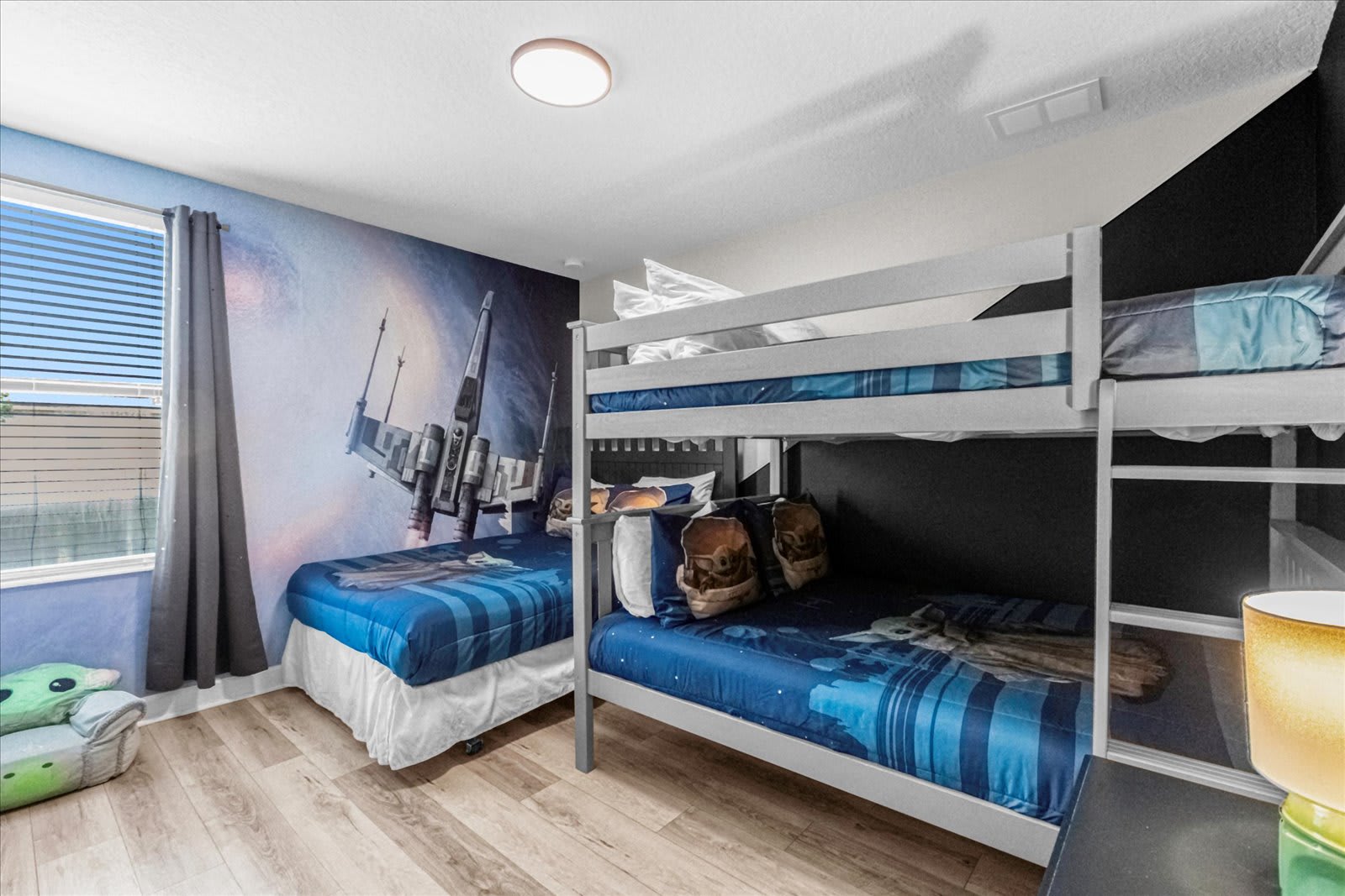 Themed bedroom featuring a large Star Wars mural on one wall, a grey bunk bed with blue striped bedding, and a single bed with matching blue bedding, all on light wood-look flooring.