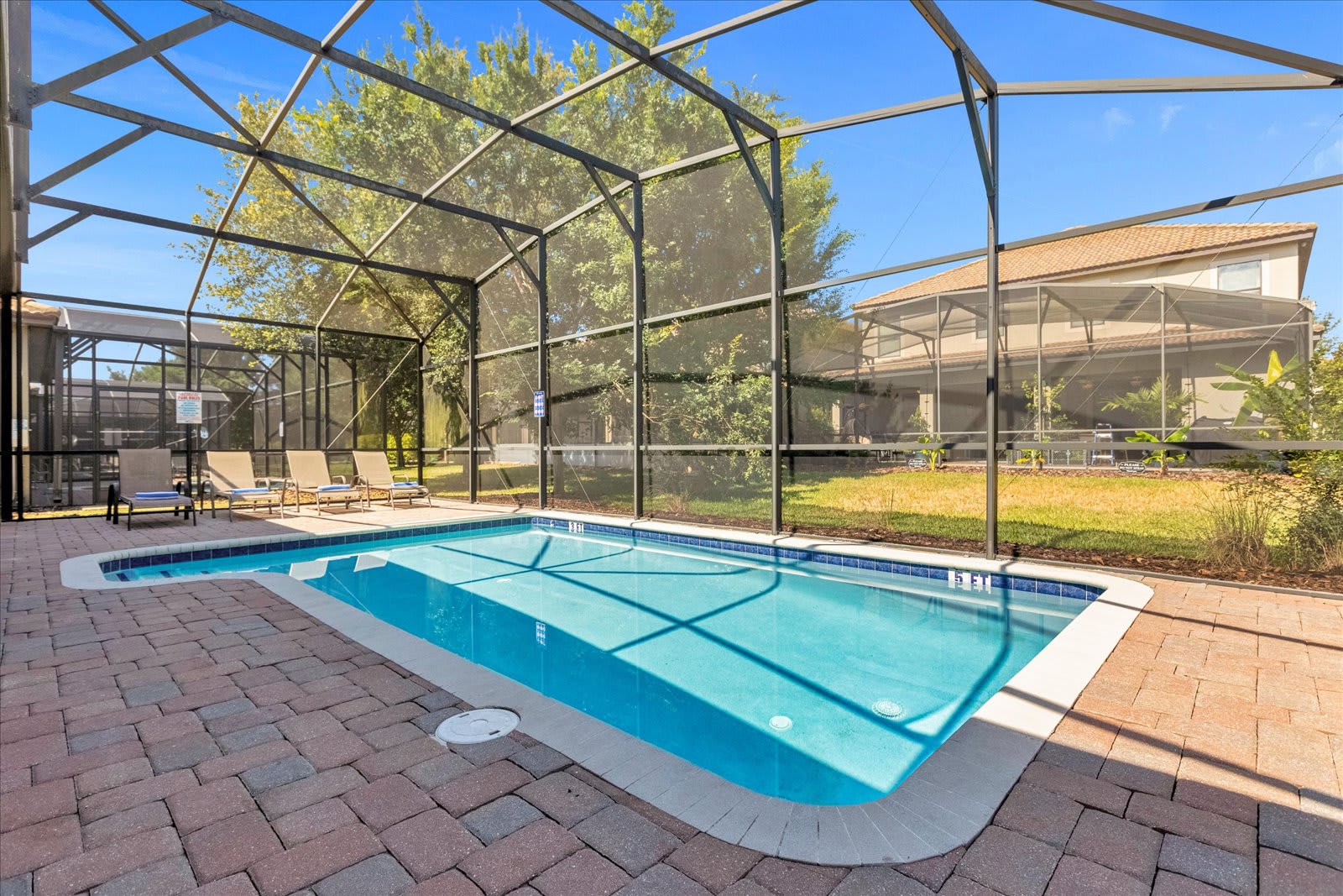 A private rectangular swimming pool with blue water, surrounded by brick pavers and enclosed by a screen cage. Several lounge chairs are visible on the pool deck, with a grassy area and other houses in the background.