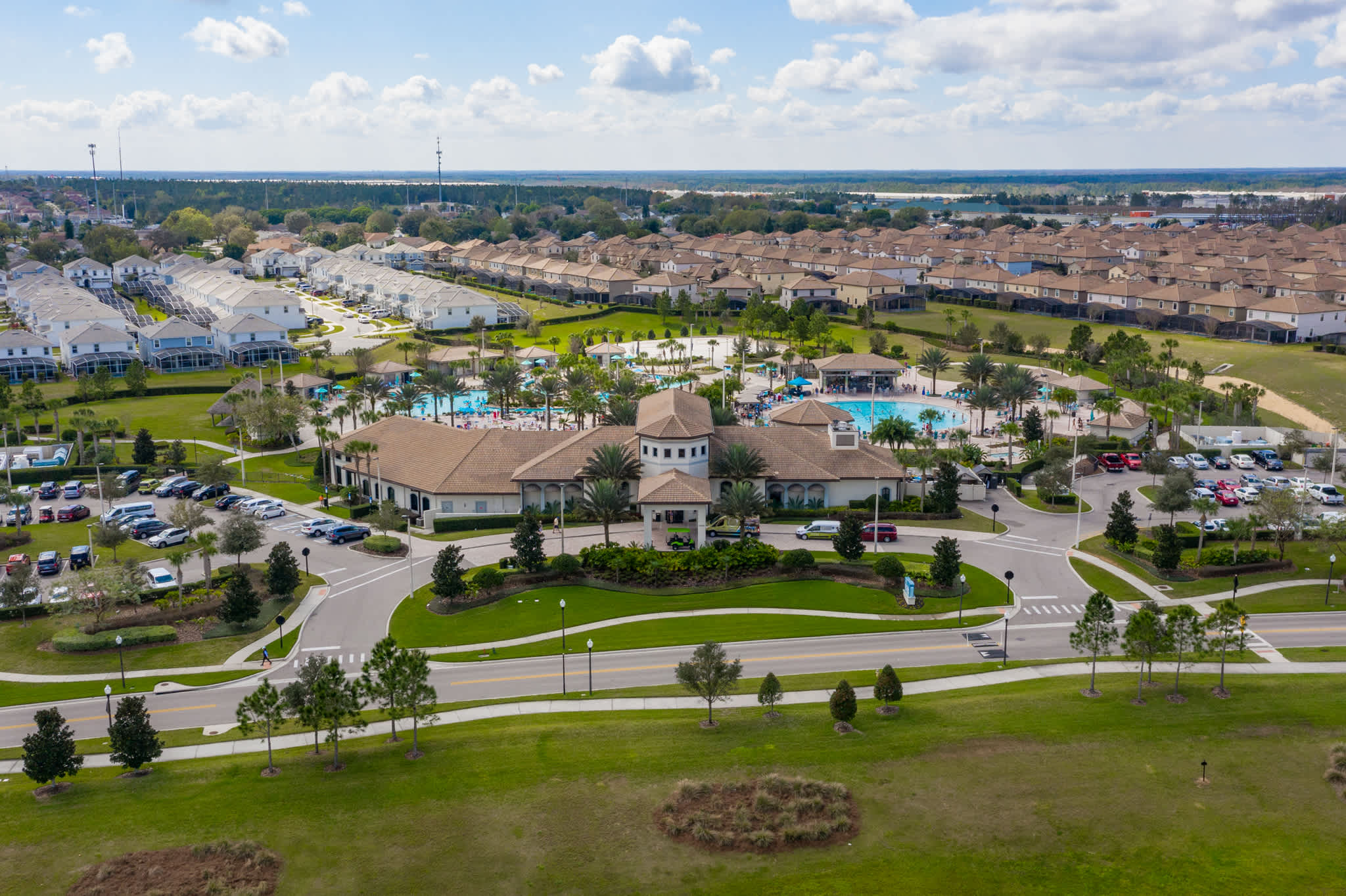 Aerial view of a large resort community featuring a central clubhouse with multiple swimming pools, surrounded by numerous residential townhouses and green spaces under a clear sky.