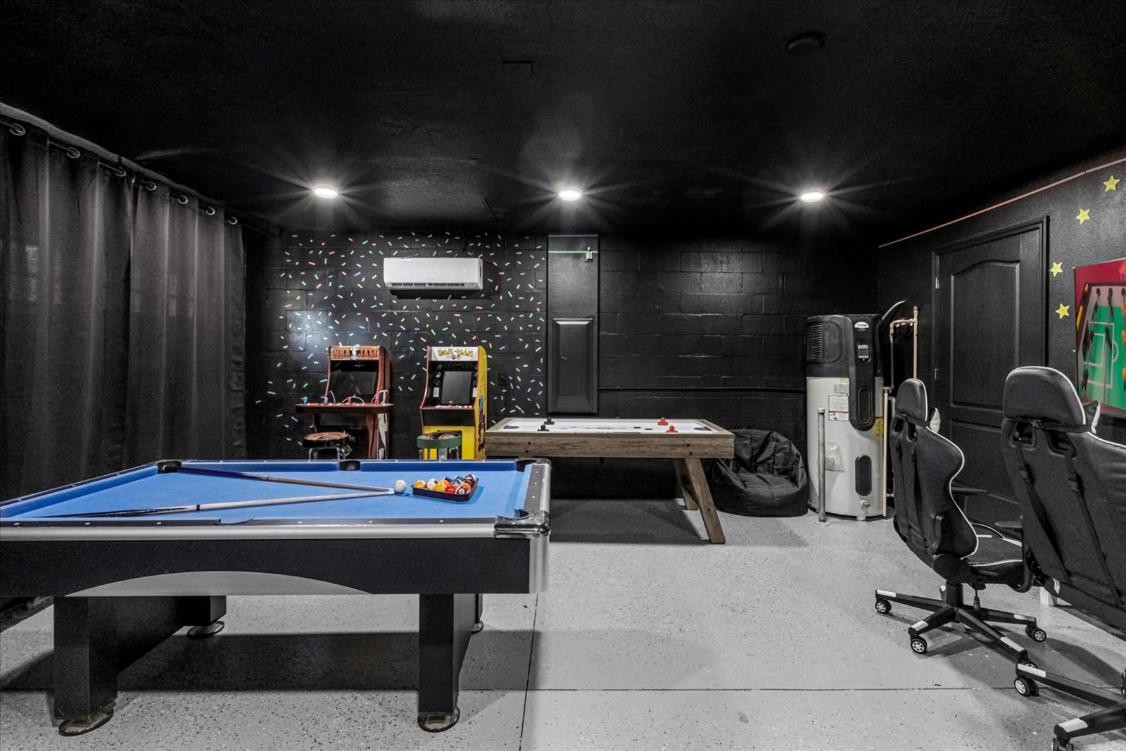 Another view of the dark-themed game room, showcasing a pool table with blue felt, two arcade machines, and a foosball table against a wall with a colorful geometric pattern. Gaming chairs are visible in the foreground.