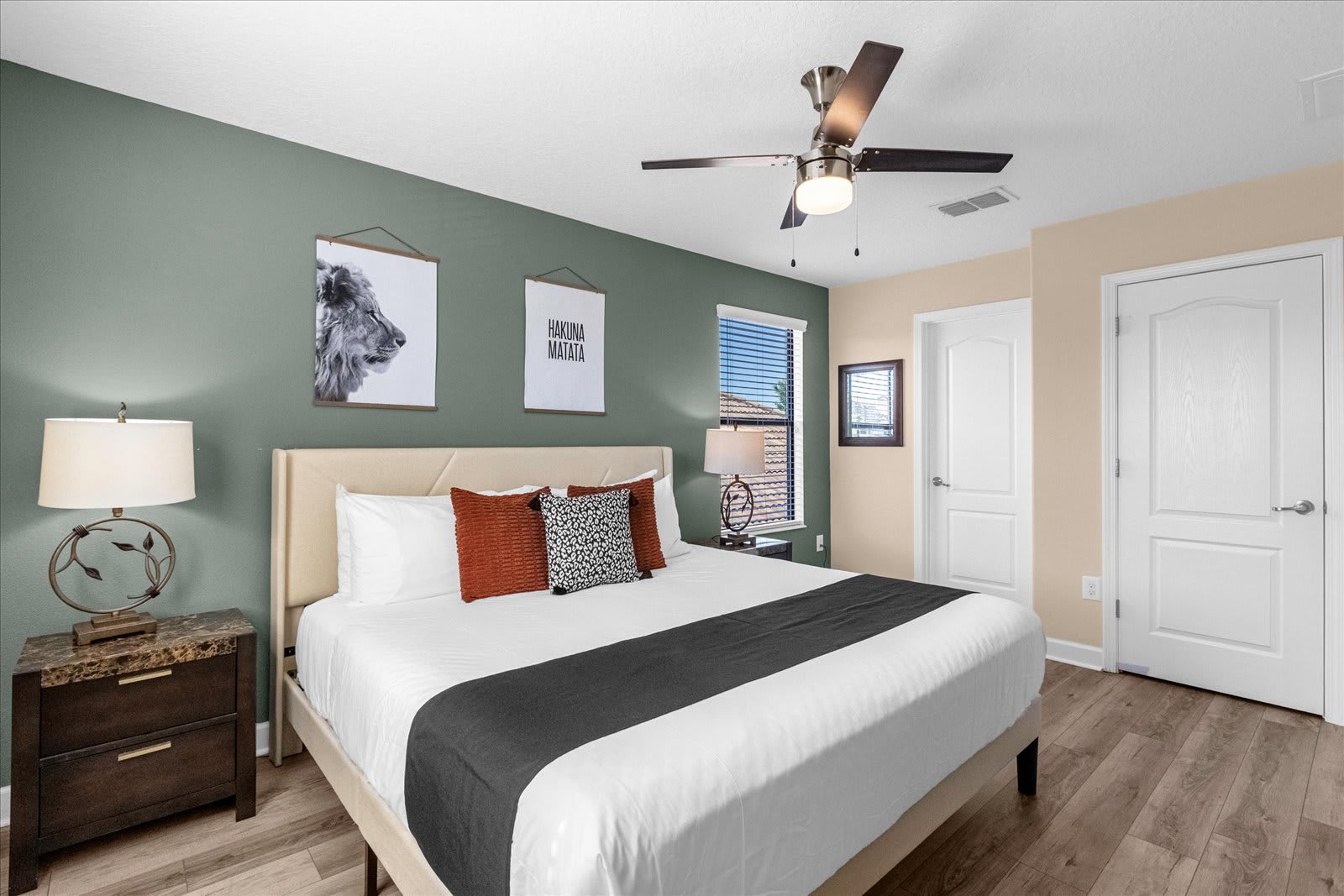 Cozy bedroom featuring a king-sized bed with a light upholstered headboard, white bedding, and decorative pillows. The room has a sage green accent wall with framed art, light wood-look flooring, and two dark nightstands with elegant lamps.