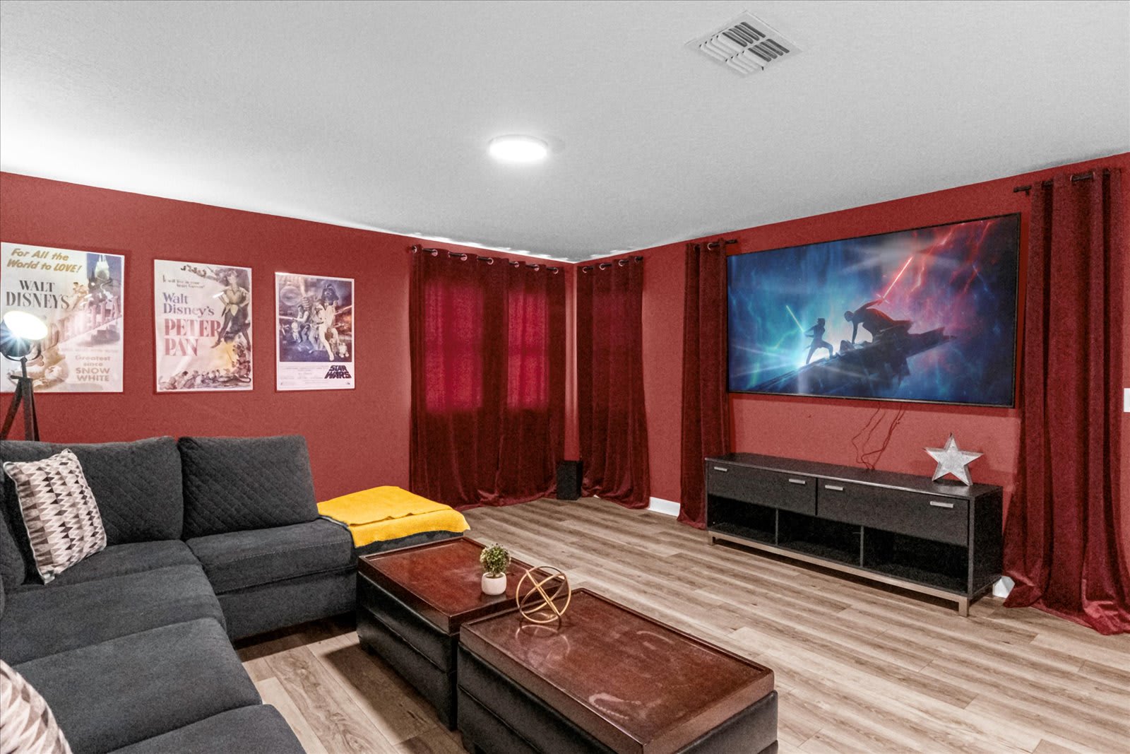 Cozy media room with a large wall-mounted TV, a dark sectional sofa, and two ottoman-style coffee tables. The room features red walls, red curtains, and movie posters, creating a cinematic ambiance.