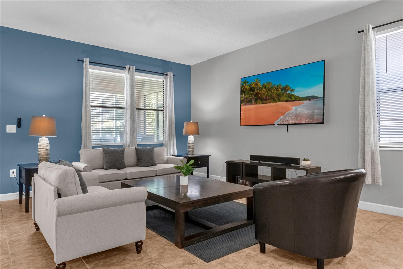 A bright living room with a blue accent wall and light gray walls, featuring a large flat-screen TV mounted above a media console. The room includes a light-colored sofa, an armchair, and a dark wood coffee table on a gray rug, with tiled floors throughout.