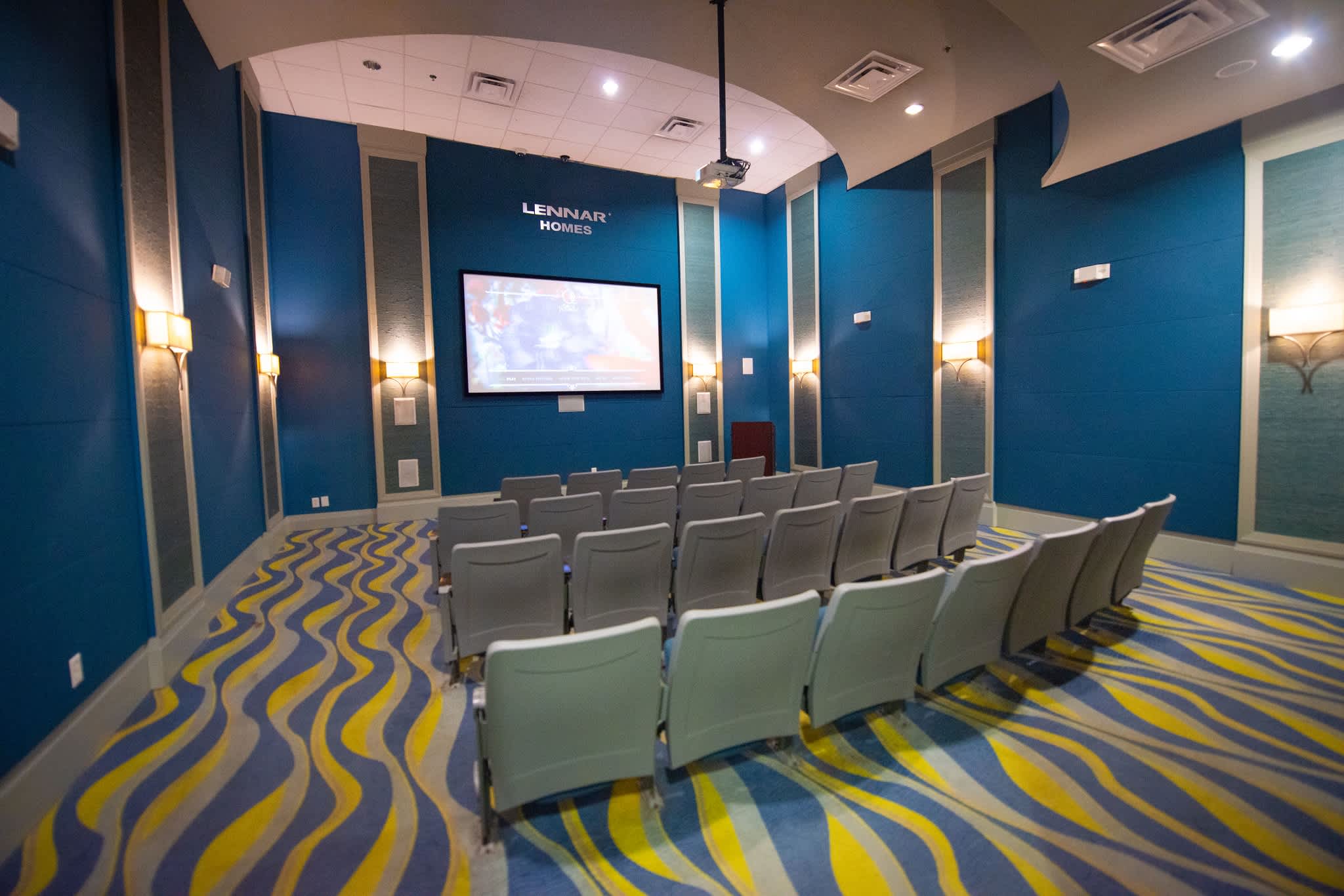 A dedicated media room with rows of grey theater-style seating facing a large wall-mounted screen, set against vibrant blue walls with decorative panels and a distinctive wavy blue and yellow carpet.