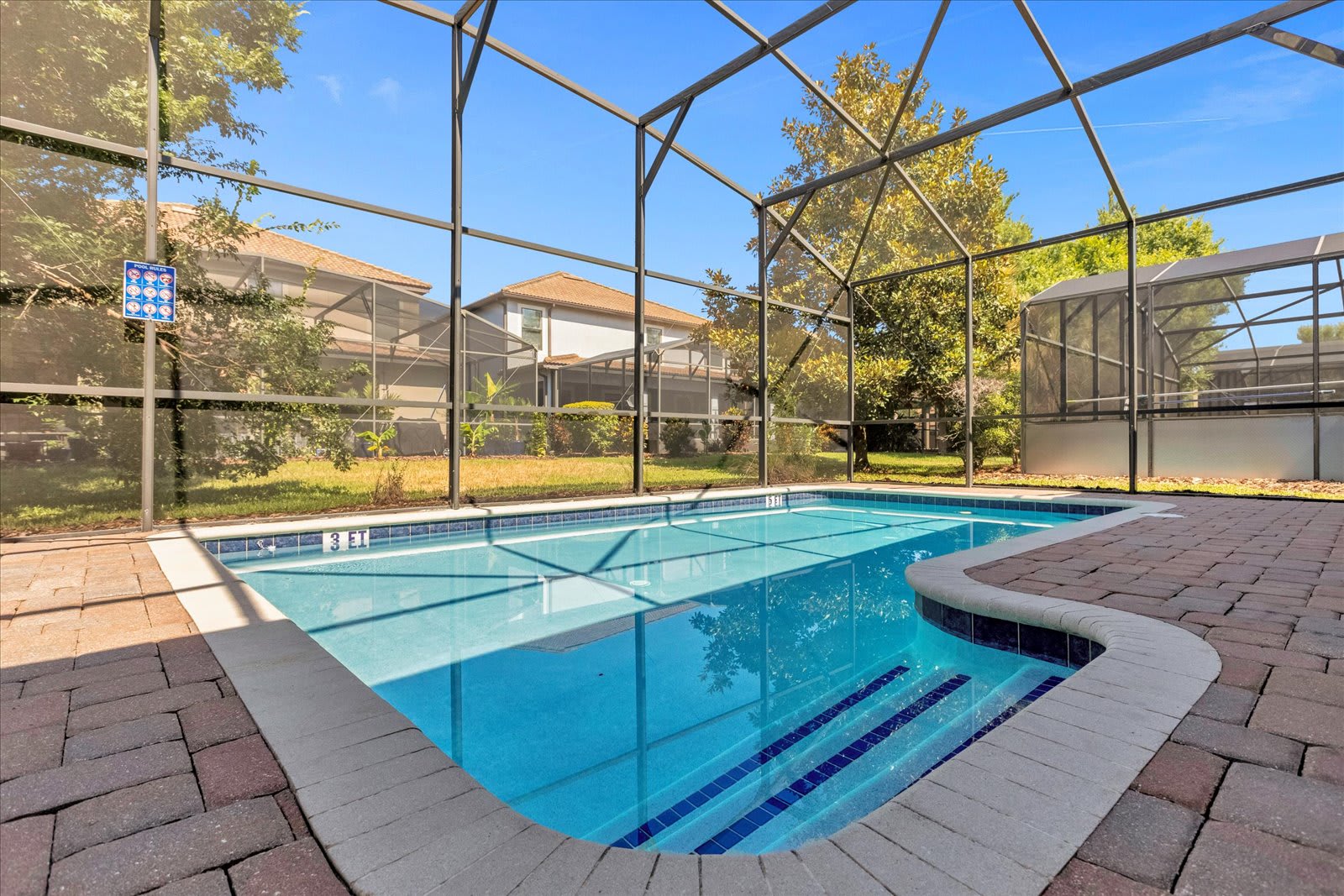 A close-up view of a private swimming pool with blue water and tiled steps, surrounded by brick pavers and a screen enclosure. The pool area is sunny, with a grassy backyard visible beyond the screen.