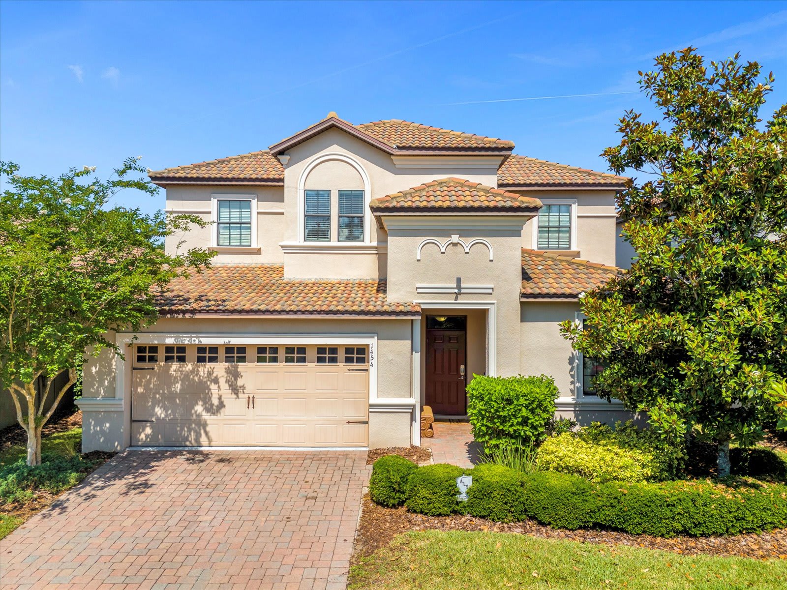 The front exterior of a two-story house with a Mediterranean-style tiled roof and light stucco walls, featuring a two-car garage and a paved driveway. Lush green landscaping with shrubs and trees frames the entrance.