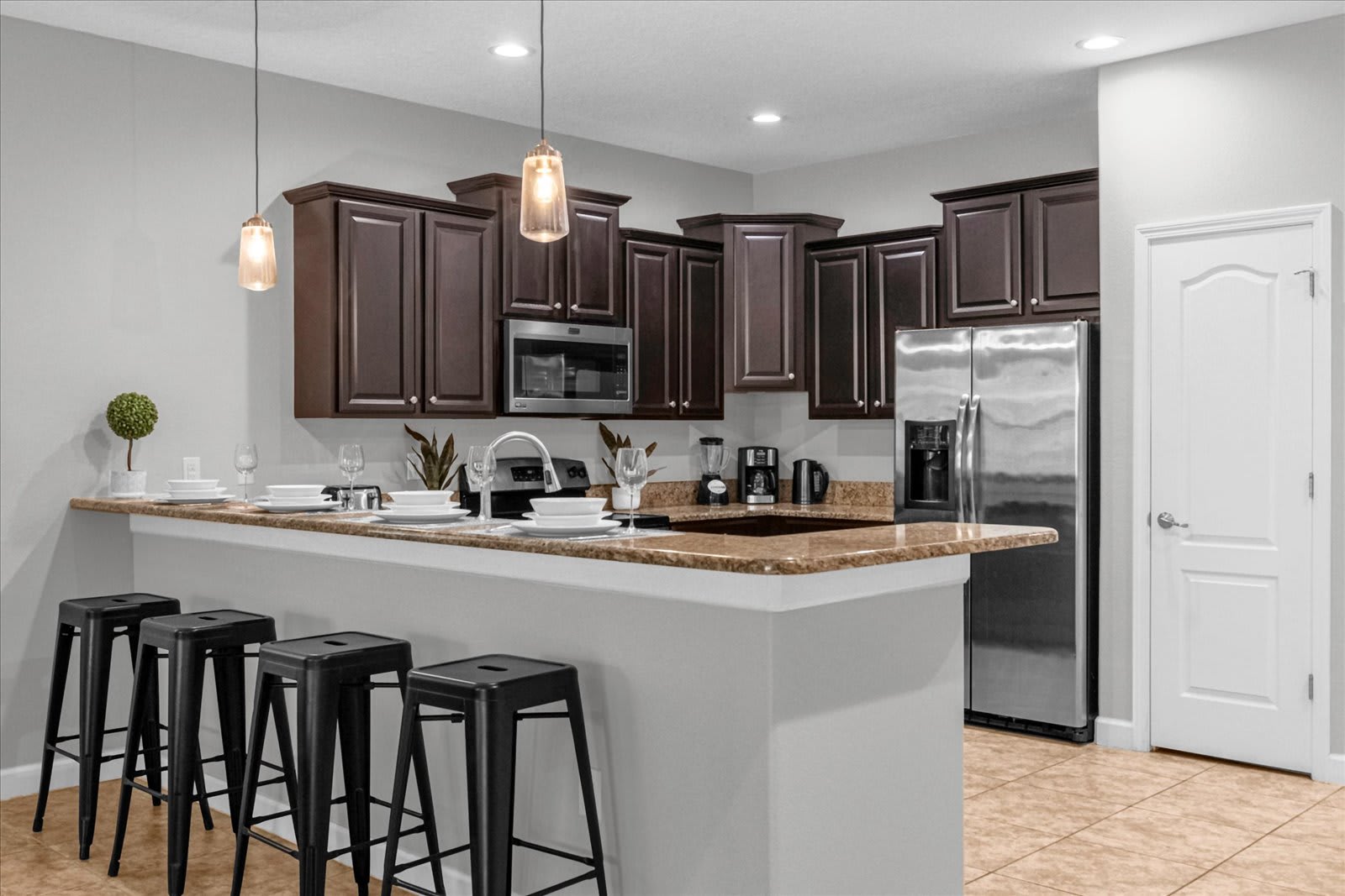Modern kitchen bar area with dark wood cabinets, granite countertops, and four black bar stools. Pendant lights hang above the bar, which is set with dishes, and a stainless steel refrigerator is visible on the right.