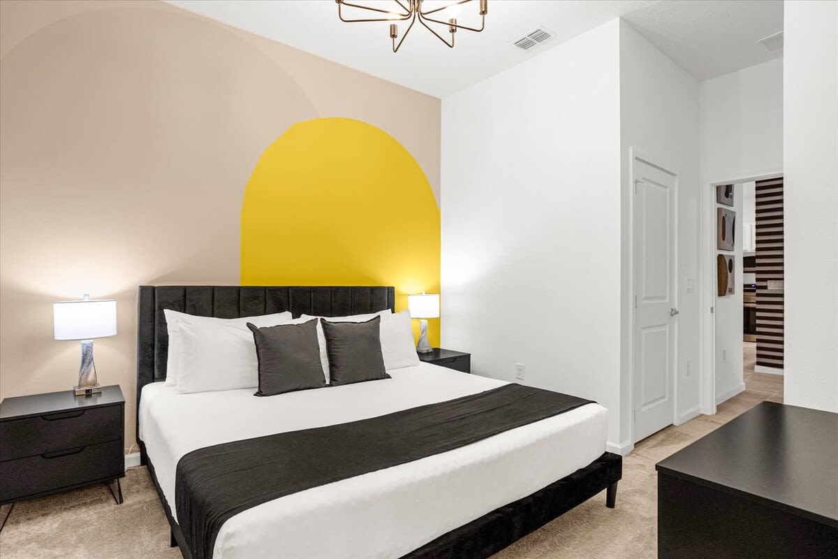 Contemporary bedroom showcasing a large bed with a dark headboard, black nightstands, and a striking accent wall with a prominent yellow circle design. A modern chandelier illuminates the space.