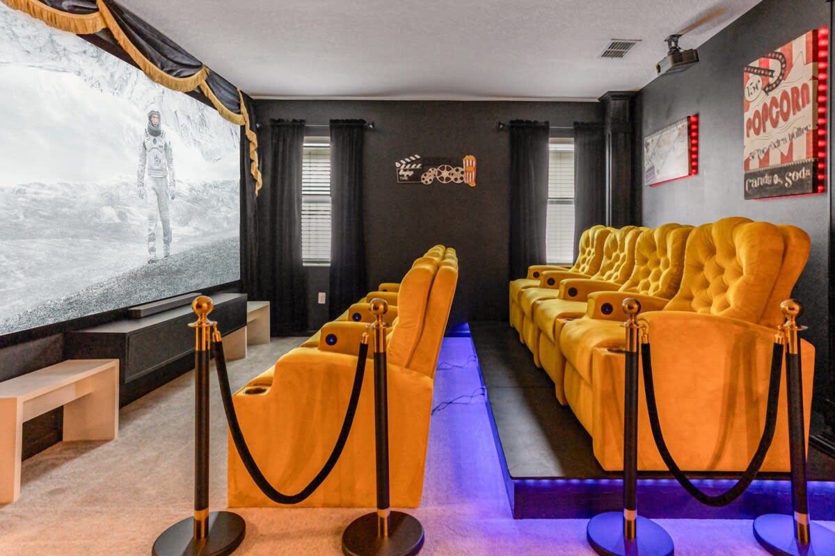 A luxurious home theater room with two tiered rows of plush yellow velvet recliners, a large projection screen displaying a black and white movie scene, and red velvet ropes for a classic cinema feel.