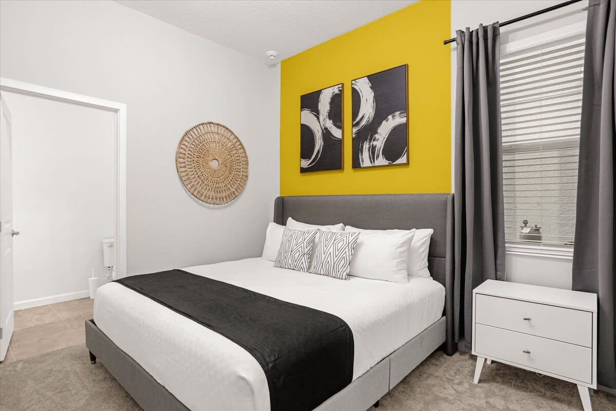 Modern bedroom with a king-sized bed, a gray upholstered headboard, and a striking yellow accent wall behind the bed, featuring two black and white abstract art pieces.