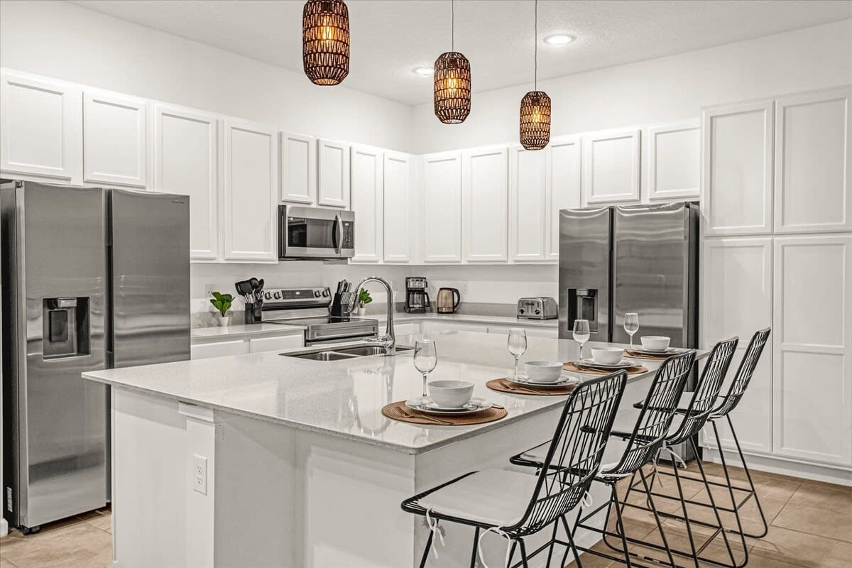 A bright, modern kitchen features white shaker-style cabinets, light quartz countertops, and stainless steel appliances including a double-door refrigerator, microwave, and range. A large island with a sink and dishwasher is visible, with three woven pendant lights overhead.