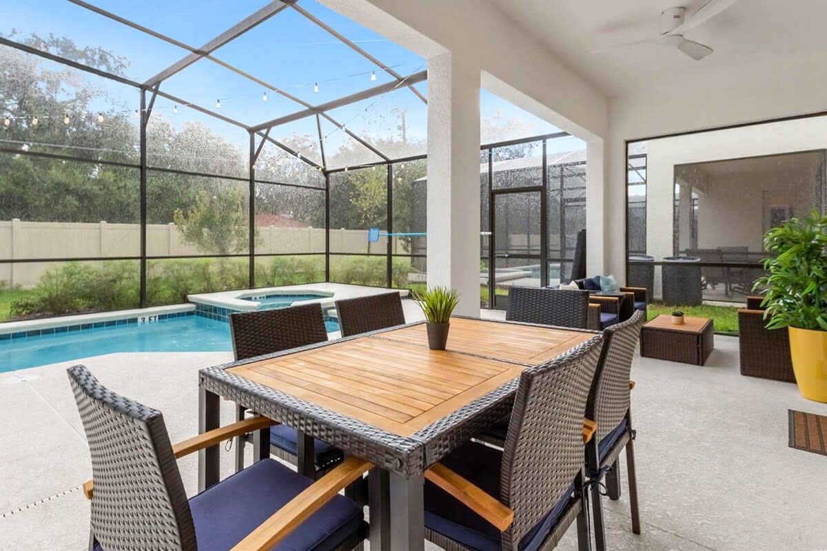 A spacious covered patio features a wooden-top dining table with dark wicker chairs and a separate lounge area, all overlooking the screened private pool. The area is well-lit and includes a ceiling fan for comfort.