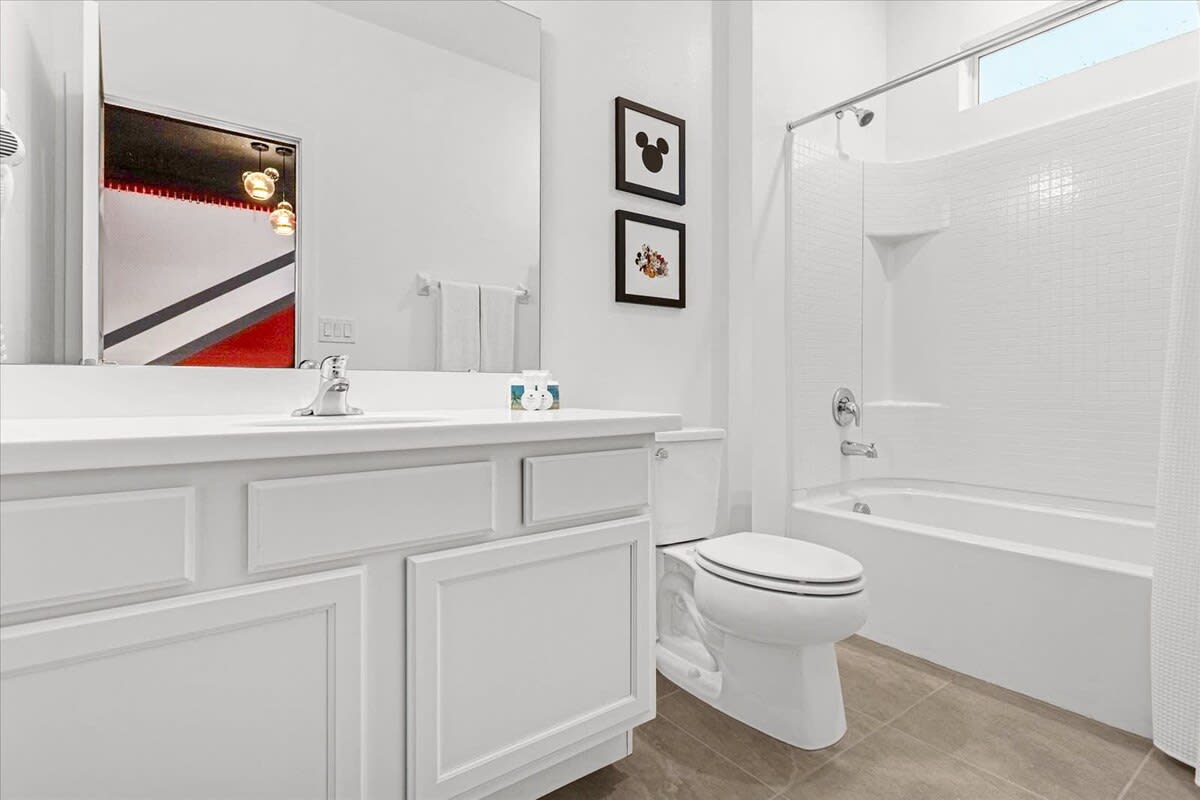 A clean white bathroom with a single vanity, a toilet, and a shower-bathtub combo. The mirror reflects the black and white Mickey Mouse themed bedroom, and two framed Mickey Mouse silhouette pictures adorn the wall.