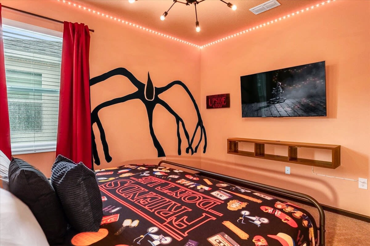 An orange-walled bedroom with a full-size bed featuring a Stranger Things themed comforter, a large wall decal of the Mind Flayer, and a wall-mounted TV. Red curtains and LED strip lights complete the themed decor.