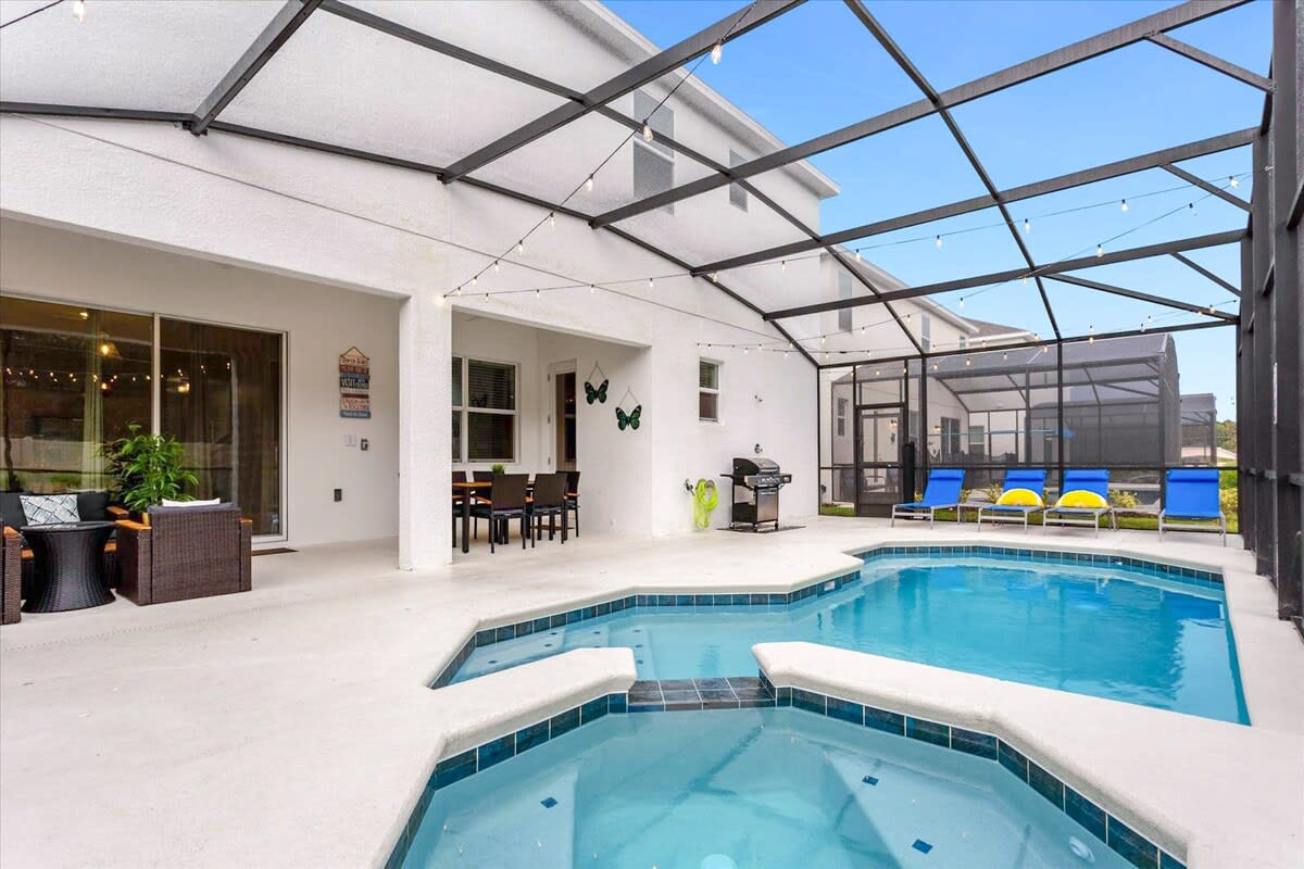 A spacious screened-in patio features a blue-tiled pool with an integrated hot tub, a covered dining area with a dark wicker table and chairs, and a separate lounge area with comfortable seating. String lights hang overhead, adding to the ambiance.