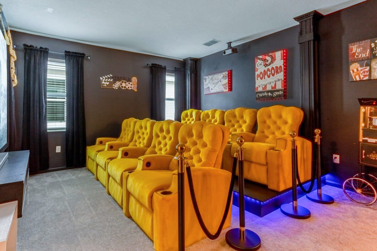 A wide view of the home theater, showcasing two rows of opulent yellow velvet recliners on a raised platform, a large screen, and movie-themed decor including popcorn signs and film reel accents.