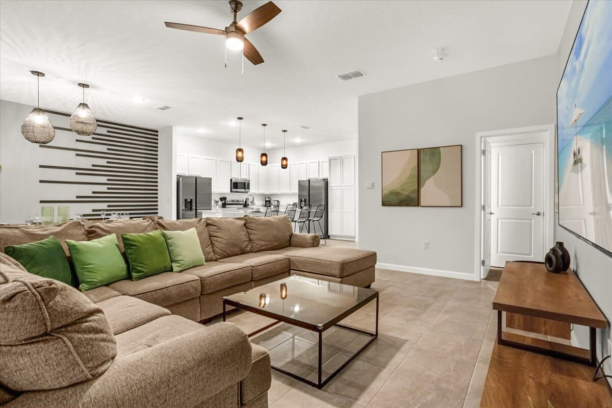 A spacious open-concept living room features a large L-shaped brown sectional sofa with green accent pillows, a glass-top coffee table, and a wall-mounted TV displaying a beach scene. The room opens to a modern white kitchen with pendant lights and a dining area.