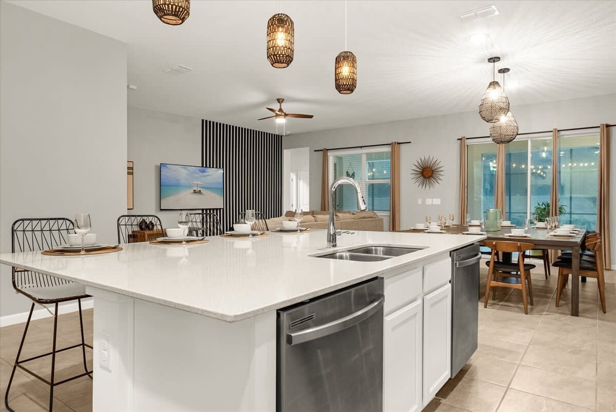 An expansive view of the open-concept living space from the kitchen island, showing the white kitchen with stainless steel appliances, a large island with a sink, and bar stools. Beyond, the dining area and living room with a large sectional sofa are visible.