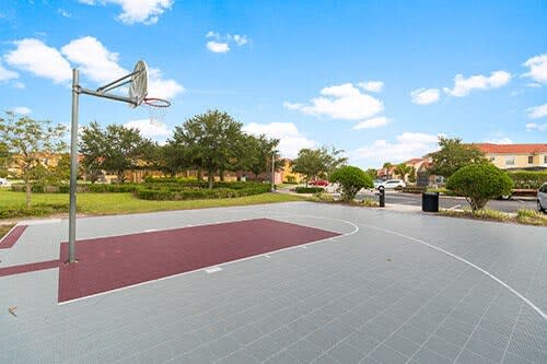 Outdoor basketball half-court with a grey and red surface and a single hoop, surrounded by green grass and trees, with residential buildings in the distance under a clear blue sky.