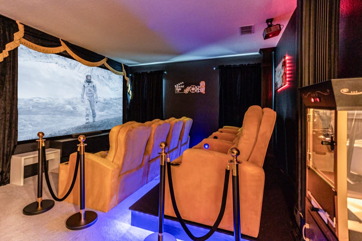 A dedicated home theater features two tiered rows of plush gold recliners, a large projection screen displaying a movie scene, and black velvet curtains. Stanchions with ropes add a classic cinema touch, and a popcorn machine is visible on the right.
