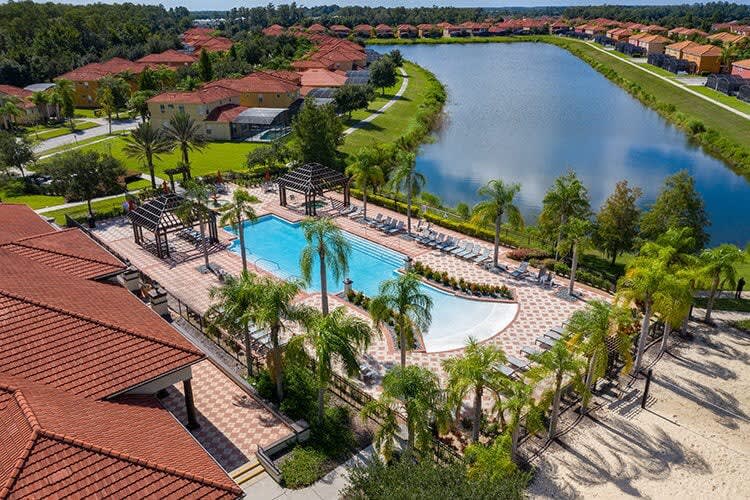Overhead aerial view of a large communal swimming pool with a shallow entry area, surrounded by lounge chairs, palm trees, and two wooden pergolas, adjacent to a large lake and residential homes.