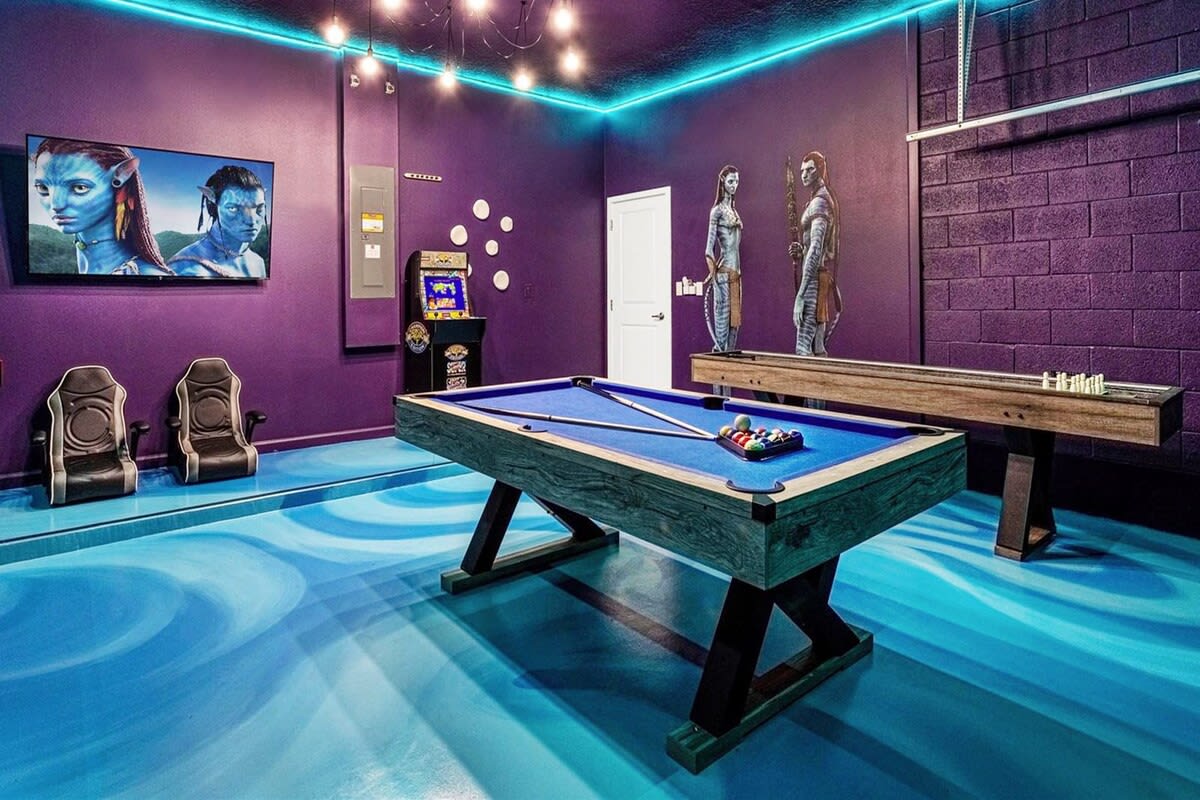 A vibrant game room with purple walls and a blue swirled floor, featuring a pool table, shuffleboard, an arcade machine, and a large TV displaying Avatar characters, all illuminated by blue LED strip lighting.