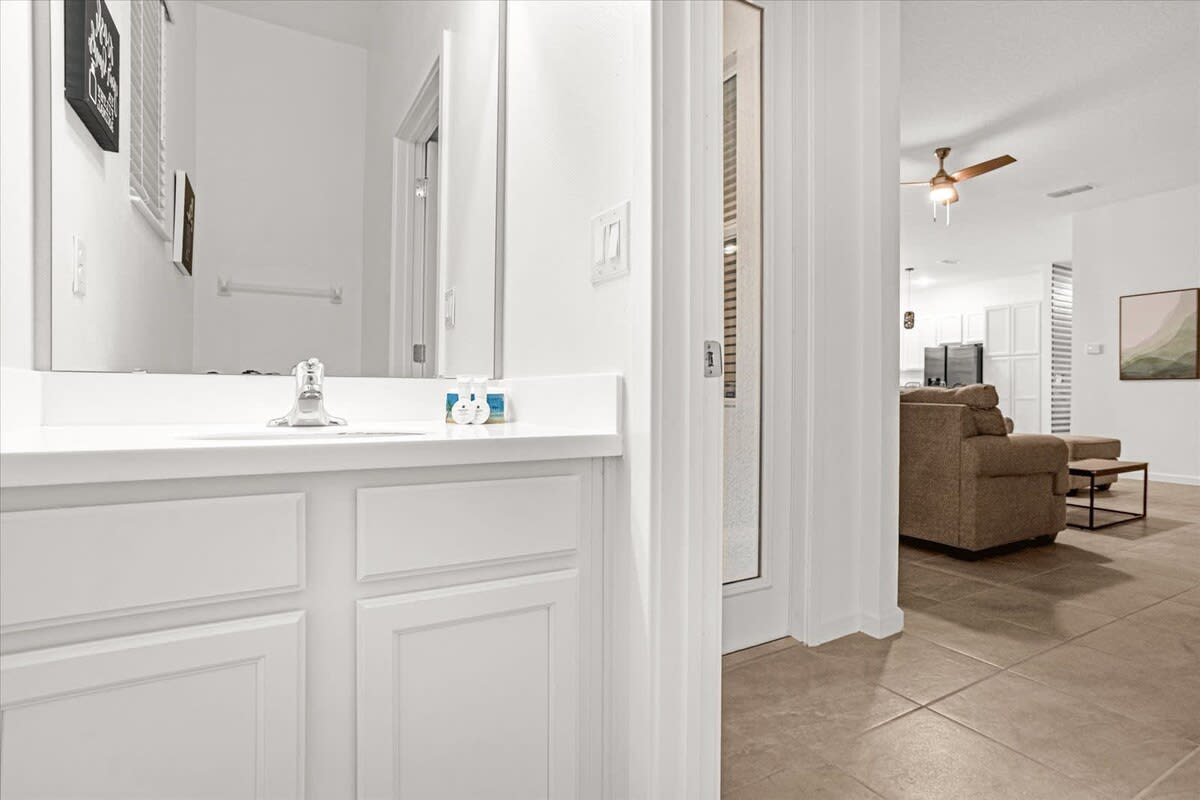 White bathroom with a single vanity and mirror, offering a partial view into an adjacent living area with a sofa and tiled flooring.