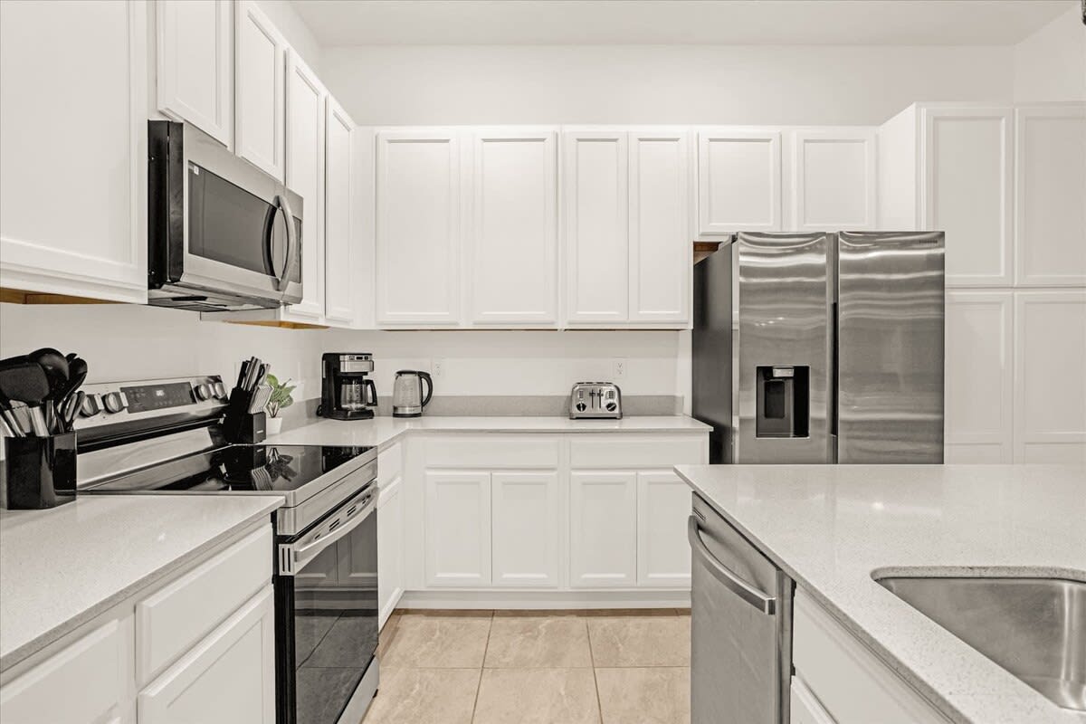 A close-up of the modern kitchen shows white shaker-style cabinets, light quartz countertops, and stainless steel appliances including a range, microwave, and coffee maker. The tile floor and a toaster are also visible.