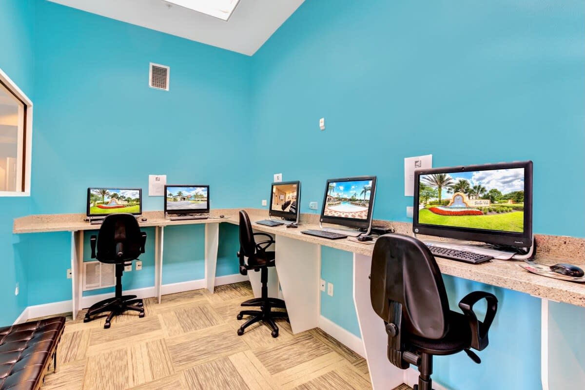 Modern business center with bright blue walls, featuring multiple computer workstations with black office chairs, light-colored countertops, and a patterned carpet, ideal for remote work or study.