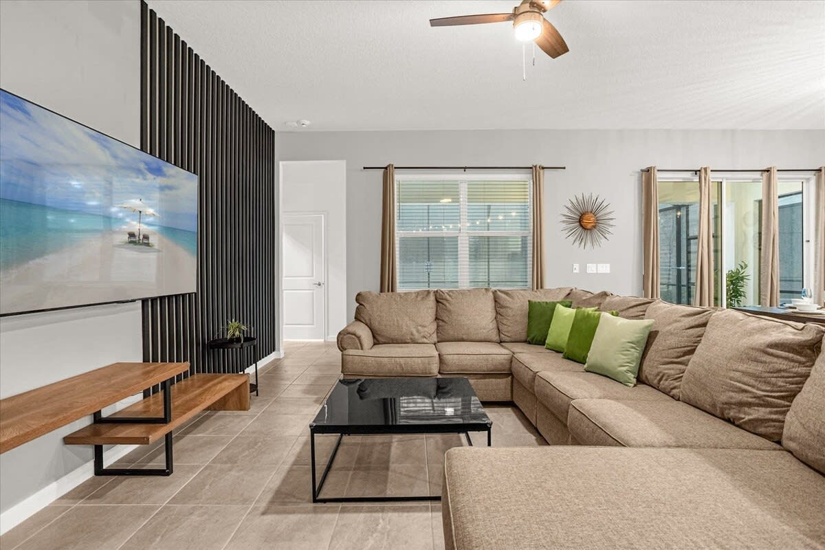 This living room features a large L-shaped brown sectional sofa with green accent pillows, a glass-top coffee table, and a large wall-mounted TV against a black slatted accent wall. A window with blinds and curtains is visible, along with a sunburst wall decor.