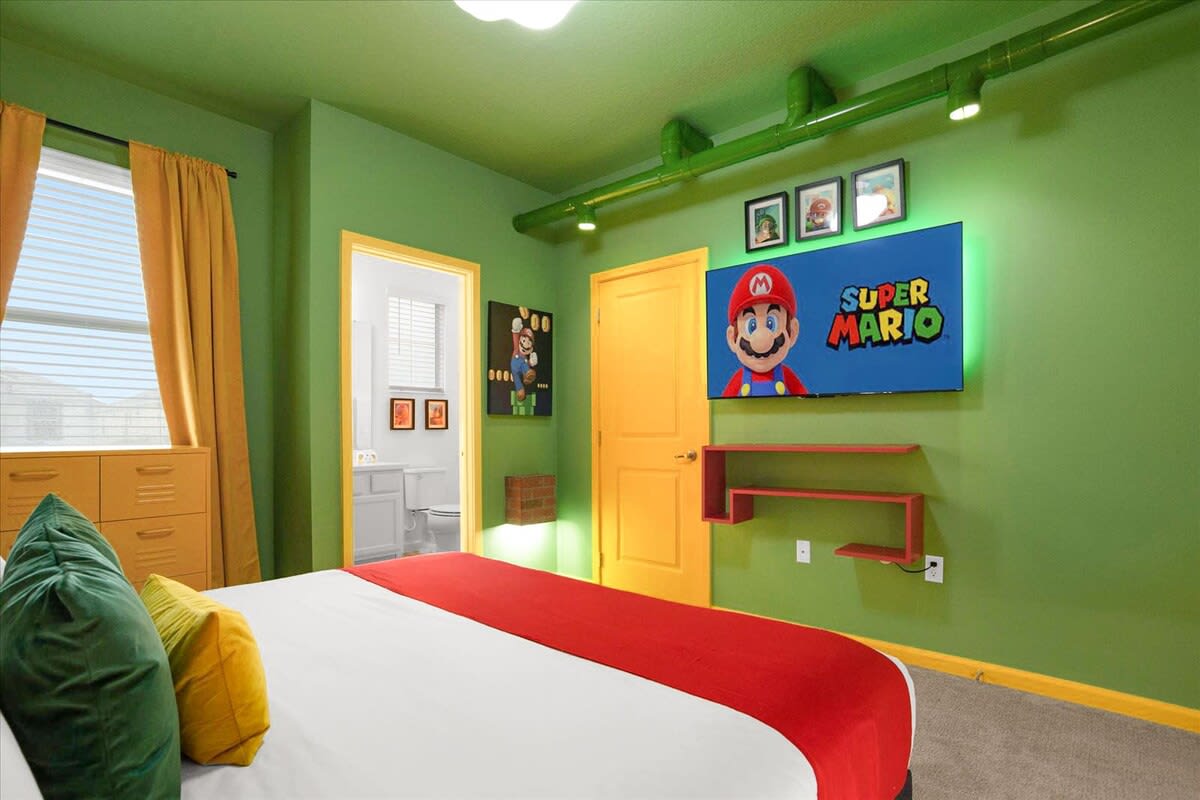 Vibrant Super Mario themed bedroom with green walls and ceiling, a queen-sized bed with red and white bedding, and a wall-mounted TV displaying the Super Mario logo.