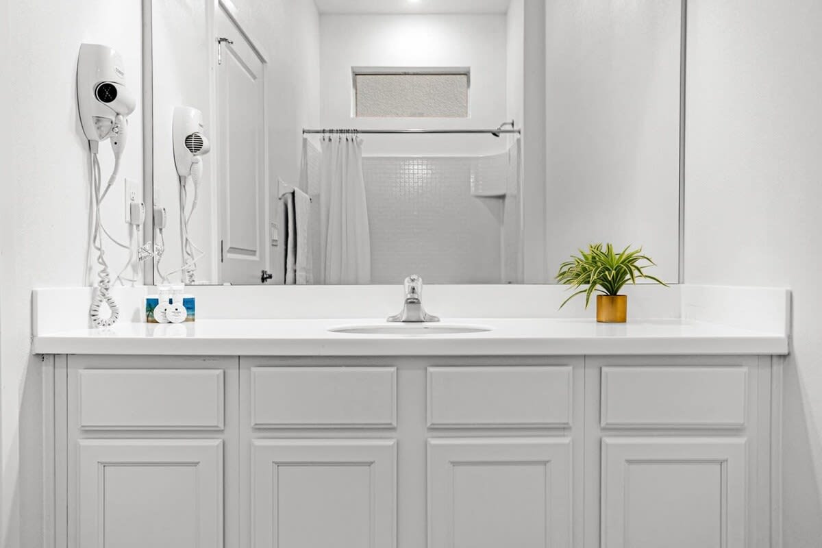 Bright white bathroom featuring a single vanity with a white countertop, a large mirror, and a shower/tub combo with a white tiled surround and shower curtain.