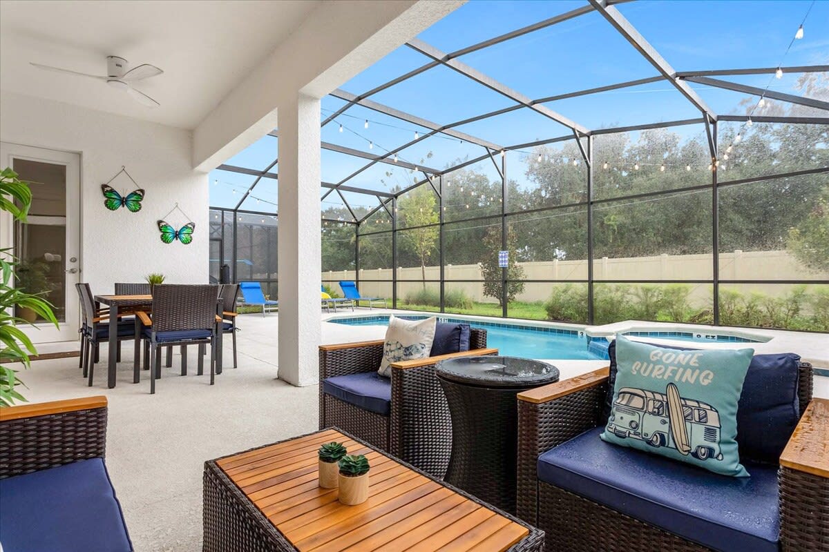A covered outdoor patio offers a comfortable lounge area with dark wicker chairs and blue cushions, alongside a dining table with matching chairs. The space overlooks the screened private pool and features a ceiling fan and decorative butterfly wall art.