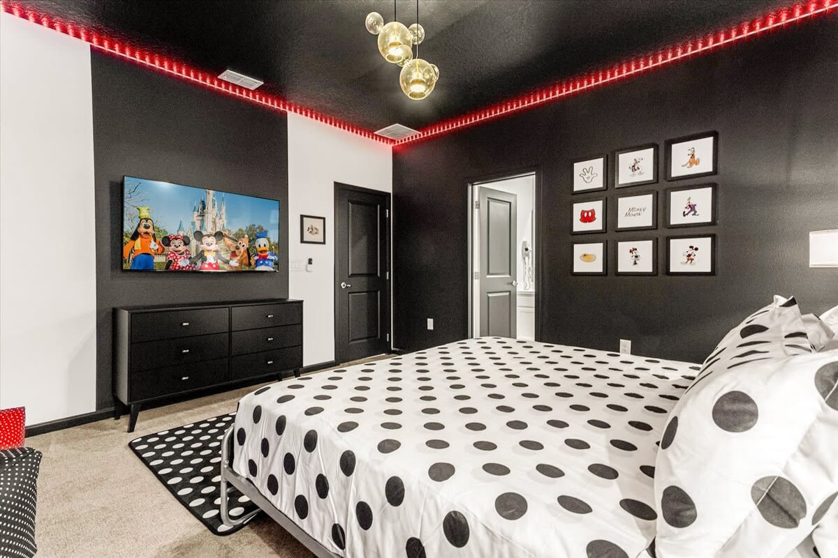 A striking black and white themed bedroom with a large bed featuring a polka-dot comforter, a black dresser with a TV, and framed Mickey Mouse artwork on the wall. Red LED strip lights line the ceiling, and a Mickey-shaped light fixture hangs above.