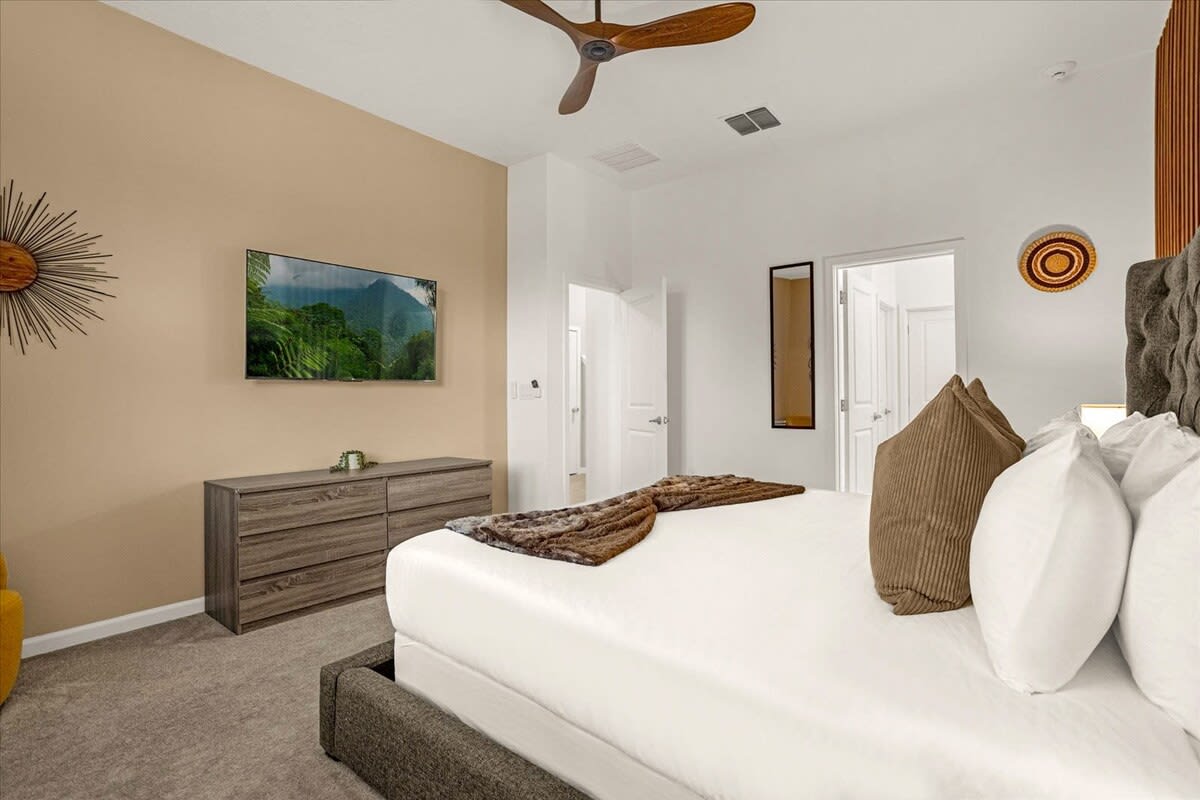 Modern bedroom with a large bed, a wall-mounted TV, and a wooden dresser against a two-tone wall (beige and white). A wooden ceiling fan hangs above.