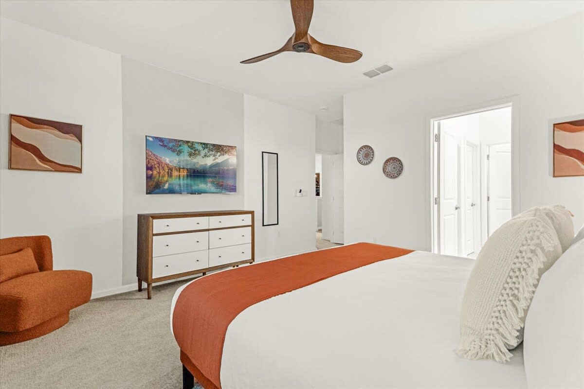 Spacious bedroom with a large bed, an orange throw, and a wall-mounted TV above a mid-century modern dresser. An orange accent chair is in the corner.