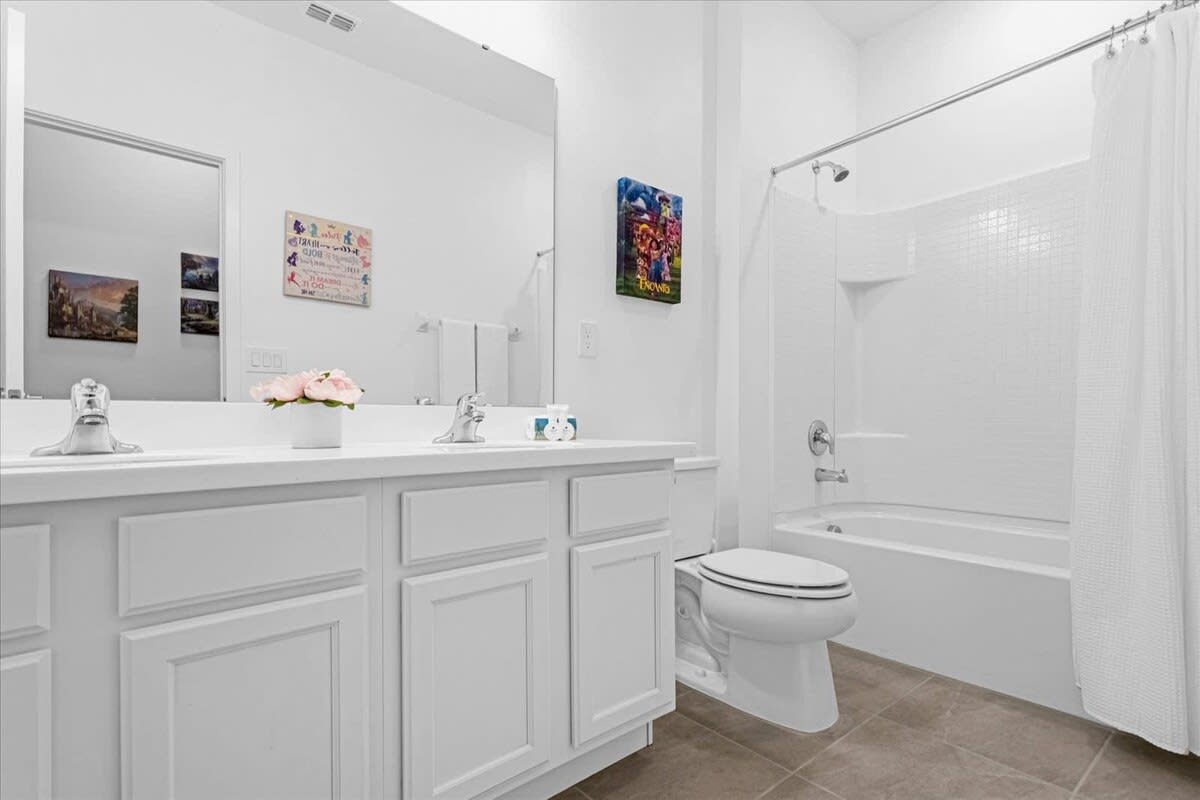 A spacious white bathroom featuring a double vanity with two sinks, a toilet, and a shower-bathtub combo. A large mirror spans the width of the vanity, and a small floral arrangement adds a touch of color.