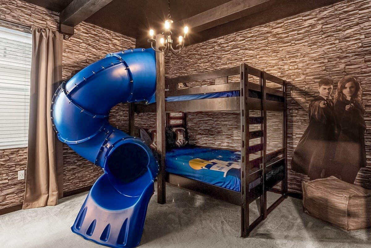 A themed kids' bedroom featuring a sturdy wooden bunk bed with a blue slide attached, set against a stone-patterned wallpaper and a large Harry Potter character cutout.