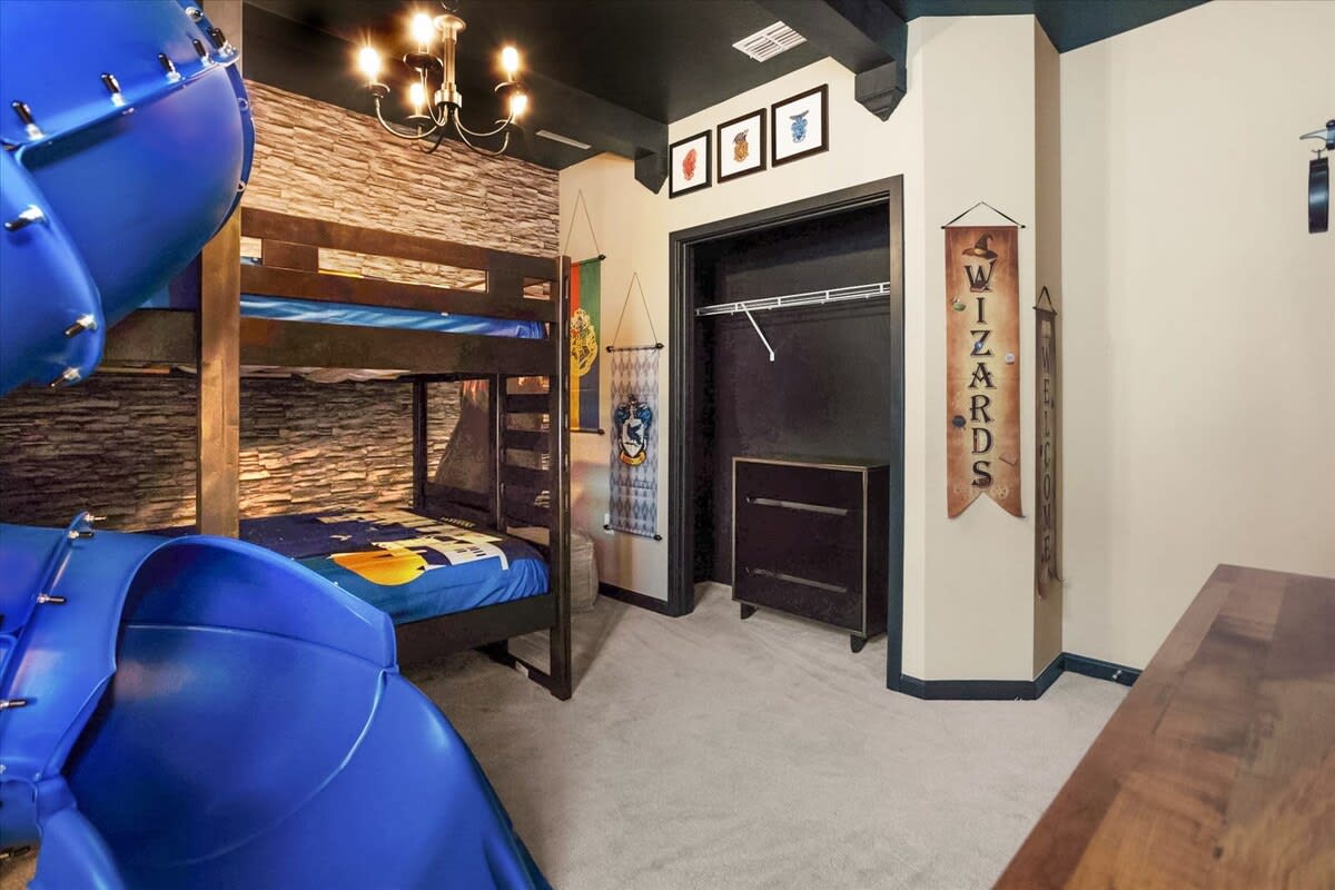 Dark and cozy Harry Potter themed bedroom featuring bunk beds with a blue slide, a stone-look accent wall, and various wizarding banners and decor.