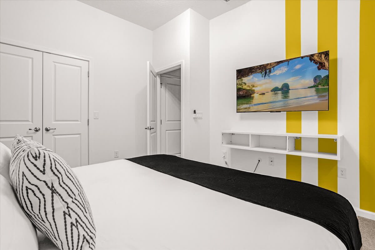 Modern bedroom with a wall-mounted TV displaying a beach scene, set against a white wall with vertical yellow stripes, and a white floating media console below.