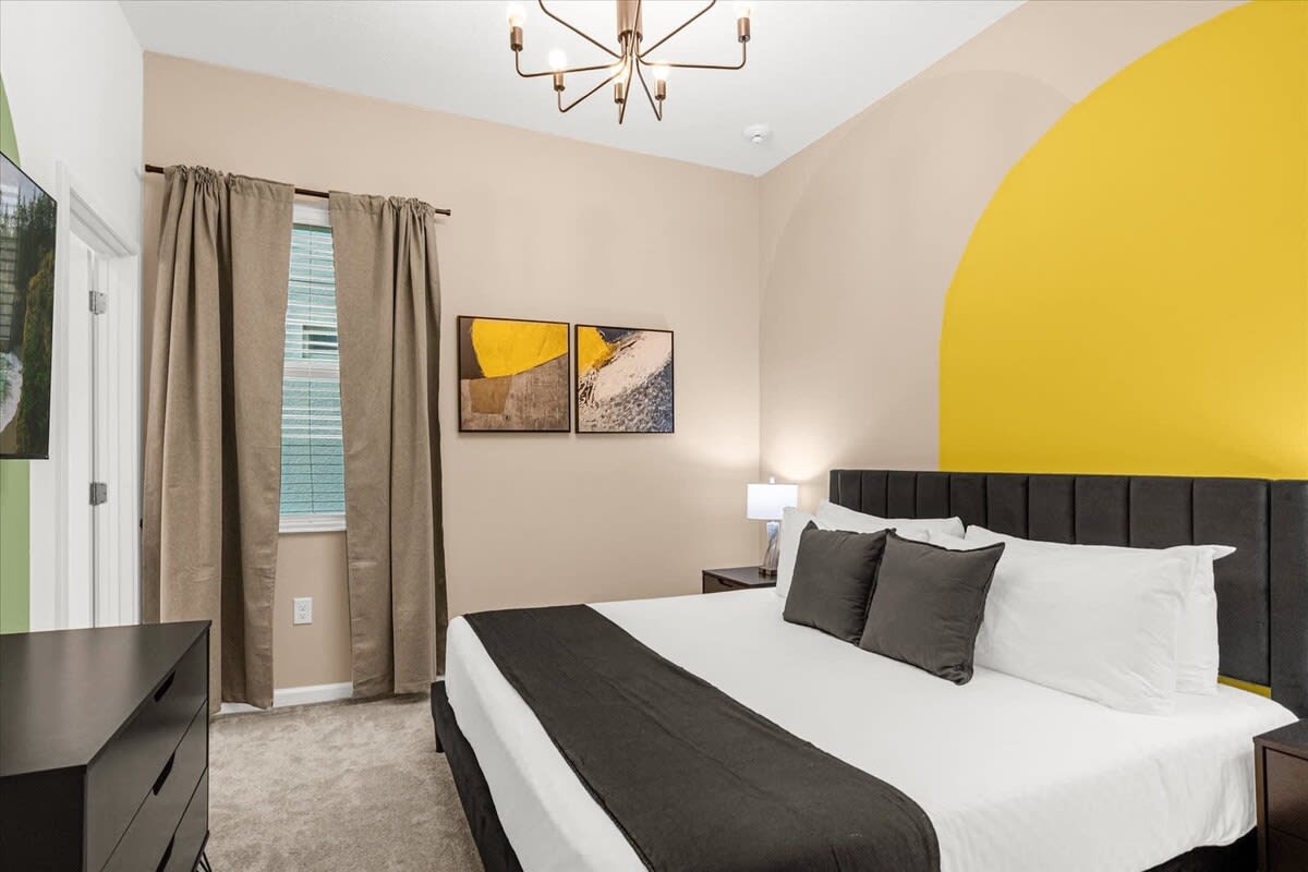 Stylish bedroom with a large bed, dark gray bedding, and a bold accent wall featuring a large yellow circle and a beige arch. A modern chandelier hangs from the ceiling.