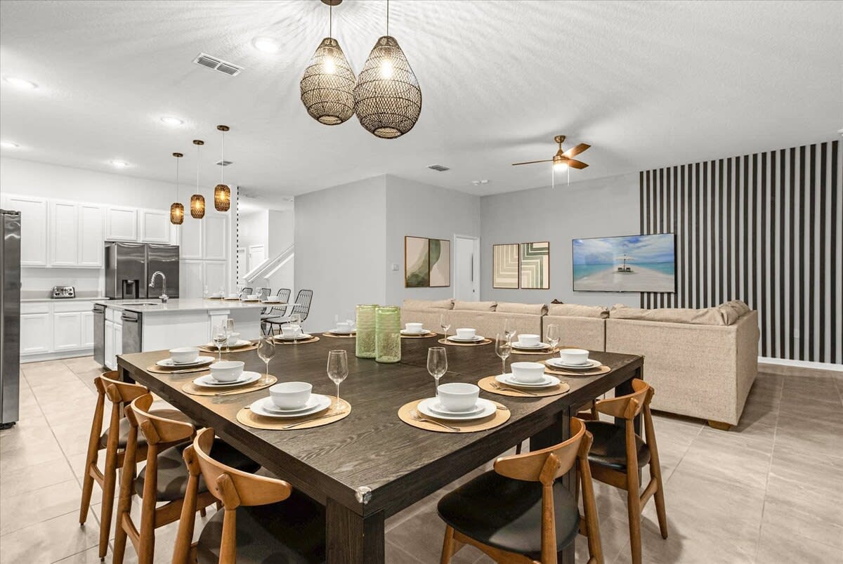 A modern open-concept space features a large square dark wood dining table with seating for eight, illuminated by two woven pendant lights. Beyond, a white kitchen with an island and a living area with a large sectional sofa and wall-mounted TV are visible.