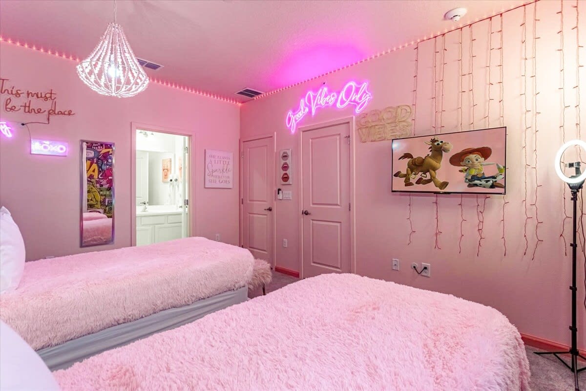A vibrant pink-themed bedroom featuring two beds with fluffy pink comforters, neon signs on the walls, string lights, and a large TV displaying an animated movie. A decorative chandelier hangs from the ceiling.