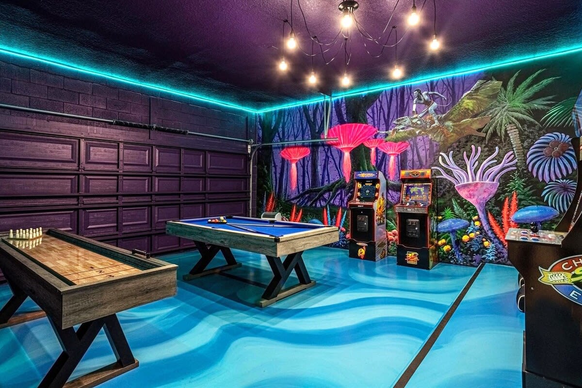 A vibrant game room features a blue-felt pool table, a shuffleboard table, and multiple arcade machines, all set against a colorful, fantastical mural. The floor is painted with blue wavy patterns, and neon strip lighting illuminates the space.