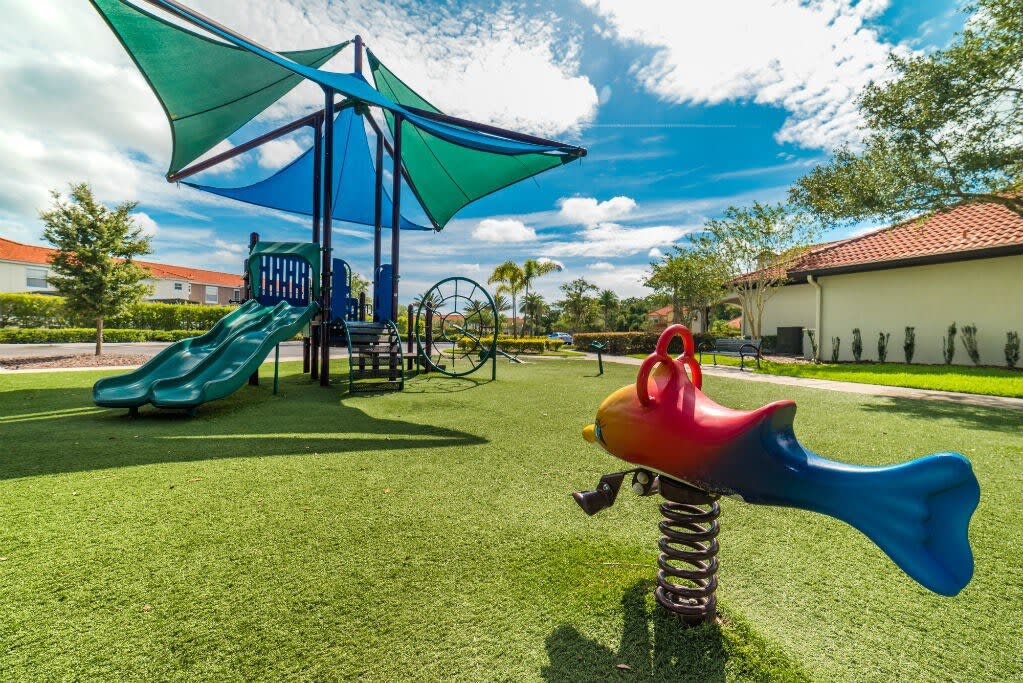 Vibrant children's playground featuring a slide, climbing structures, and a spring rider shaped like a colorful fish, all set on soft green artificial turf under a blue and green shade canopy.