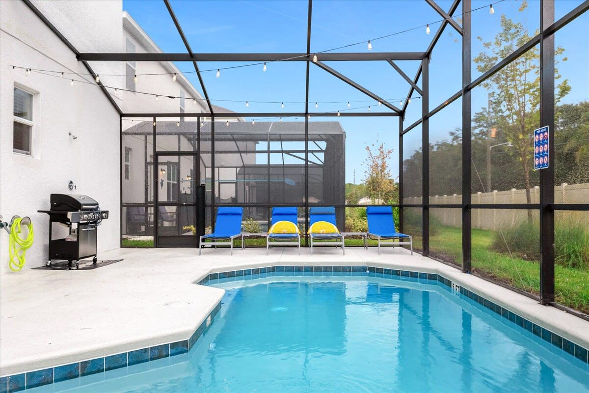 The screened-in pool area features a sparkling blue private pool with several blue and yellow lounge chairs lined up along the far edge. A BBQ grill is visible on the patio, and the screen enclosure provides a view of the surrounding fenced yard.
