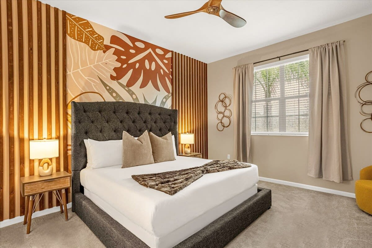 A stylish bedroom features a king-sized bed with a tufted dark gray headboard, white bedding, and a faux fur throw. The accent wall behind the bed has vertical wood slats and a large botanical mural in earthy tones, complemented by a wooden ceiling fan.