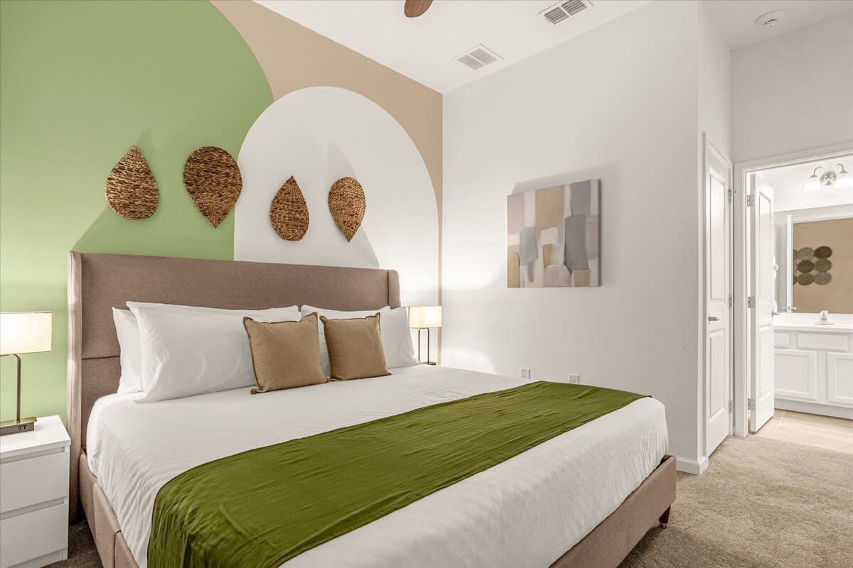 A modern bedroom features a king-sized bed with a tall upholstered headboard, white bedding, and a green throw blanket. The accent wall behind the bed has a geometric design in green, beige, and white, with three woven leaf-shaped wall decorations.
